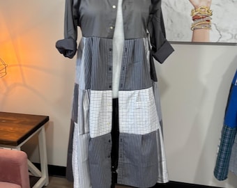 May include: A long, gray patchwork dress on a black mannequin stand. The top portion is a solid gray, button-down shirt. The skirt is made of various gray and white striped and checkered fabrics. The sleeves are rolled up.