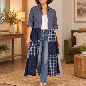 May include: A long-sleeved button-down shirt dress featuring a solid blue denim top and a patchwork skirt bottom composed of navy blue, white, and light blue fabric panels with plaid and polka dot patterns, worn over blue denim jeans.