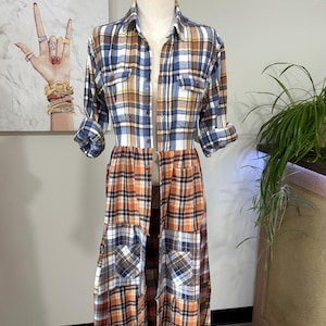 May include: A long, patchwork plaid shirt dress in shades of blue, brown, orange, and white. The dress features a collared neckline, button-down front, and long sleeves rolled up to the elbows. The skirt portion is a patchwork design.