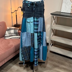 Puede incluir: Un vestido patchwork de mezclilla y cuadros en un maniquí. El vestido presenta un corpiño de mezclilla con un detalle de lazo y una falda patchwork en tonos azules y turquesas. El vestido se exhibe en un soporte negro.