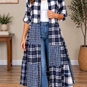 May include: A long-sleeved, button-down shirt dress featuring a navy blue and white plaid pattern. The garment has a collared neckline, two chest pockets, and a tiered, patchwork-style skirt design. It is worn over a white tank top and blue jeans.