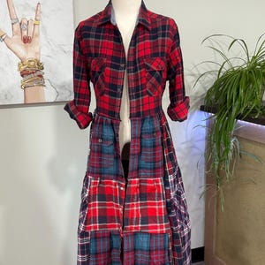 Op de afbeelding: Een patchwork geruite jurk in rood, marineblauw en blauwgroen. De top is een rood geruit button-down shirt met twee borstzakken. De rok is een patchwork van verschillende ruitpatronen. De jurk wordt op een mannequin getoond.