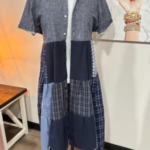 May include: A patchwork dress on a black mannequin. The top is a short-sleeved, dark gray button-down shirt over a white t-shirt. The skirt is a mix of blue and navy fabrics, including plaid and solid patterns. The dress is displayed on a black metal stand.