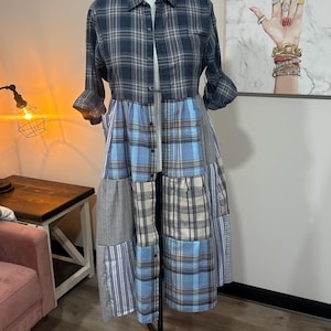 May include: A patchwork dress on a black mannequin stand. The top is a dark blue plaid shirt with rolled-up sleeves. The skirt is a patchwork of blue, grey, and white fabrics in various patterns. The dress is displayed in a room with a painting on the wall.