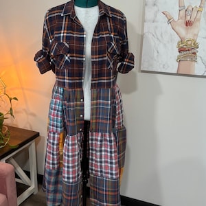 Könnte beinhalten: Ein langärmeliges Hemdblusenkleid mit einem Oberteil aus dunkelbraunem, marineblauem und weißem Schottenkaro und zwei Brusttaschen. Der Rock besteht aus einem Patchwork aus verschiedenen karierten Stoffen in Rot-, Blau-, Gelb- und Grüntönen.