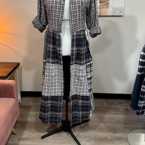 Op de afbeelding: Een lange, patchwork geruite overhemdjurk op een mannequin. Het bovenste gedeelte is een overhemd met knopen met een rood, zwart en wit geruit patroon. De onderkant is een patchwork van verschillende geruite stoffen in grijstinten, zwart en wit.