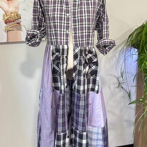 May include: A patchwork dress on a mannequin. The top is a button-down shirt with a purple, black, and white plaid pattern. The skirt is a mix of purple and plaid fabrics. The sleeves are rolled up.