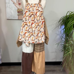 Puede incluir: Una blusa sin mangas con un patrón abstracto multicolor en tonos marrón, naranja y blanco, combinada con pantalones anchos. Los pantalones tienen un diseño de patchwork con secciones en marrón, beige, blanco y tela de cuadros marrón en la cintura.