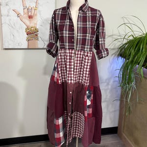 Puede incluir: Vestido patchwork sobre maniquí. El vestido tiene una parte superior abotonada con cuello y un estampado de cuadros burdeos y blancos. La falda es un patchwork de telas burdeos, rojas y blancas, incluyendo cuadros vichy y texturas lisas. El vestido está sobre un soporte plateado.