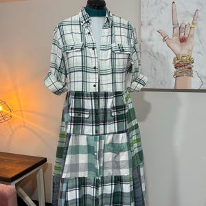 May include: A patchwork dress made from repurposed plaid shirts. The dress features a collared neckline, button-down front, and a mix of green, white, and gray plaid patterns. The dress is displayed on a dress form.