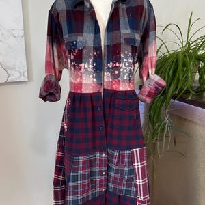 Op de afbeelding: Een lange, knoopsluitende jurk gemaakt van hergebruikte flanellen shirts. De jurk heeft een patchwork ontwerp met verschillende ruitpatronen in tinten rood, blauw en grijs. Het bovenste gedeelte heeft een gebleekt effect en de mouwen zijn opgerold.