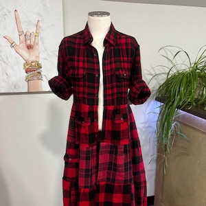 May include: A red and black plaid shirt dress on a mannequin. The dress has a collared neckline, long sleeves, and multiple pockets. The dress is a patchwork design with a tiered skirt. The dress is displayed in a well-lit room.