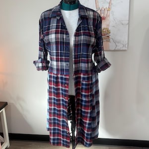 Könnte beinhalten: Ein langärmeliges, durchgeknöpftes Hemdkleid mit einem Karomuster in Marineblau, Rot und Weiß. Das Kleidungsstück verfügt über einen Kragen, eine Knopfleiste auf der Vorderseite und hochgekrempelte Ärmel und wird auf einer schwarzen Schneiderpuppe präsentiert.