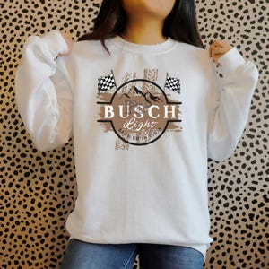 Pode incluir: Camisola branca com um gr&aacute;fico castanho de um logotipo de cerveja Busch Light com bandeiras quadriculadas e marcas de pneus. O texto no logotipo diz "Busch Light Brewed in U.S.A."
