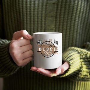 Pode incluir: Caneca de cer&acirc;mica branca com um gr&aacute;fico marrom e branco de uma bandeira quadriculada, montanhas e o texto "Busch Light Brewed in St. Louis".