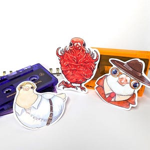 May include: A selection of bird-themed stickers. One sticker shows a red bird, another a bird in a white shirt and belt, and a third bird wearing a hat, glasses, and a suit. A purple cassette tape is also visible in the image.