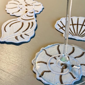 May include: A set of white fabric coasters featuring embroidered seashell designs in metallic bronze thread. The collection includes various spiral and fan-shaped shell motifs, with one coaster supporting the base of a clear glass stemware.
