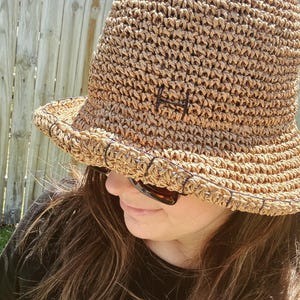 May include: A woven straw bucket hat in a natural tan color. The hat features a dark brown embroidered "H" on the front and a slightly curved brim. The hat is designed with a close weave and a dark brown trim.