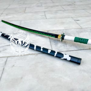 May include: A black and green katana sword with a white and green handle. The sword is lying on a white surface.