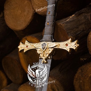 Pode incluir: Uma espada decorativa com uma empunhadura dourada e uma coroa no topo. A lâmina é feita de aço damasco e tem um padrão de linhas em espiral. A espada está apoiada em uma pilha de toras de madeira. O logotipo "Eagle Cutlery" é visível na lâmina.