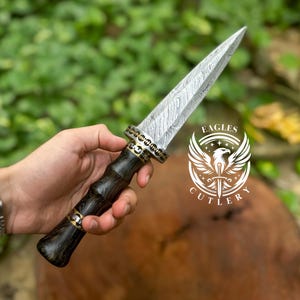 May include: A Damascus steel dagger with a black wood handle and gold accents. The blade has a distinctive pattern and is held by a hand. The background is blurred and shows green foliage. The image also includes a logo for "Eagles Cutlery" featuring a stylized eagle with a dagger in its talons.