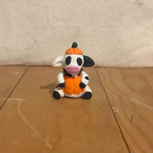May include: A handmade clay figurine of a cow wearing a pumpkin hat and vest. The cow is white with black spots and ears, a pink nose, and black eyes. The pumpkin hat and vest are orange with green accents.