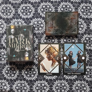 May include: A set of tarot cards with a dark background and gold accents. The box features the title "Dark Mirror Oracle of Cards". The cards depict a woman with a crown of thorns and two women standing on a staircase.
