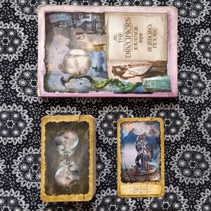 Puede incluir: Dos cartas de tarot, una con la representación de dos caras y la otra con una figura de pie sobre una roca, con el texto "The Fool" en la parte inferior. Las cartas están sobre una superficie con un patrón blanco y negro. El libro del fondo se titula "The Dreamer's Journal" de Barbara Moore.