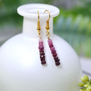 May include: A pair of dangle earrings featuring gold-toned hooks and a vertical strand of faceted beads in a gradient of pink, magenta, and deep burgundy tones. The beads are arranged from lightest at the top to darkest at the bottom.
