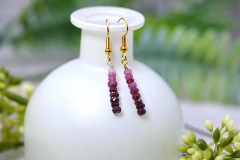 May include: A pair of dangle earrings featuring gold-toned hooks and a vertical strand of faceted beads in a gradient of pink, magenta, and deep burgundy tones. The beads are arranged from lightest at the top to darkest at the bottom.