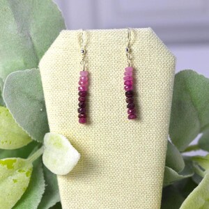 May include: A pair of drop earrings featuring a vertical row of faceted gemstone beads in a gradient colour scheme, transitioning from light pink at the top to deep magenta and dark red at the bottom, suspended from silver-toned metal hooks.