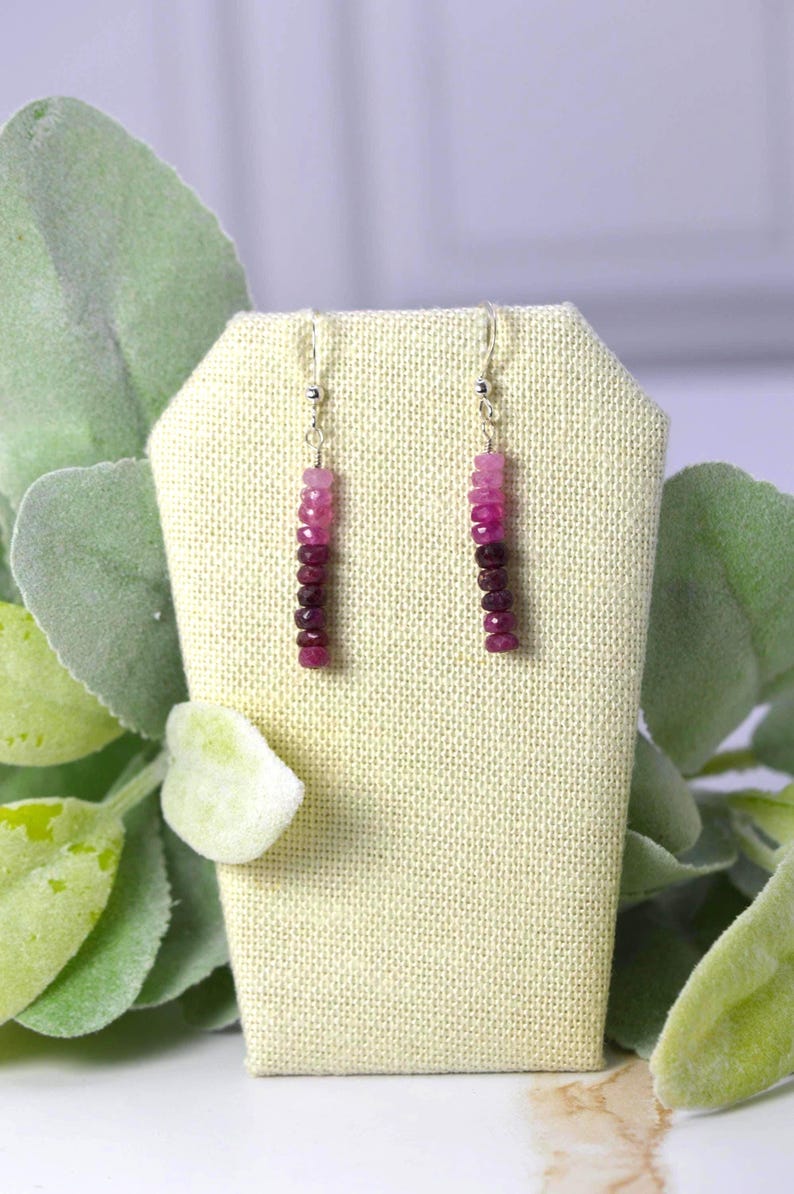May include: A pair of drop earrings featuring a vertical row of faceted gemstone beads in a gradient colour scheme, transitioning from light pink at the top to deep magenta and dark red at the bottom, suspended from silver-toned metal hooks.
