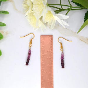 May include: A pair of dangle earrings featuring small, round, gradient-coloured pink and deep red beads on gold-toned hooks, positioned next to a copper-coloured ruler measuring approximately 5 cm in length for scale reference.