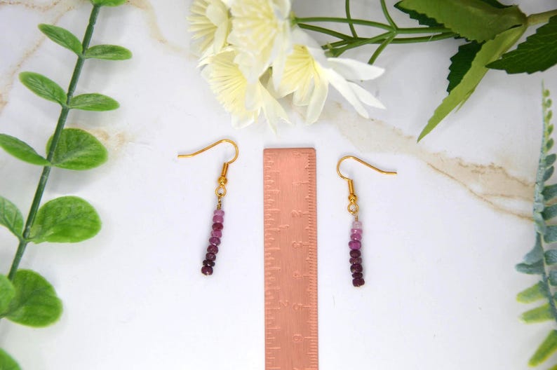May include: A pair of dangle earrings featuring small, round, gradient-coloured pink and deep red beads on gold-toned hooks, positioned next to a copper-coloured ruler measuring approximately 5 cm in length for scale reference.