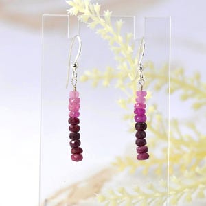 May include: A pair of drop earrings featuring a vertical row of small, faceted beads in a gradient of pink to deep burgundy red. The beads are suspended from silver-toned metal hooks, creating a linear design that hangs straight down from the top.