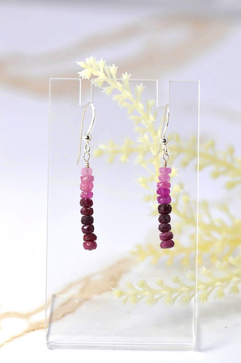 May include: A pair of drop earrings featuring a vertical row of small, faceted beads in a gradient of pink to deep burgundy red. The beads are suspended from silver-toned metal hooks, creating a linear design that hangs straight down from the top.