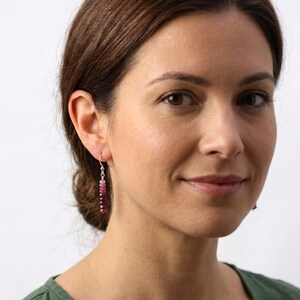 May include: A drop earring featuring a vertical row of small, faceted beads in a gradient of pink and deep magenta hues. The beads are strung on a thin silver-toned wire with a simple hook closure, creating a delicate and slender dangling design.