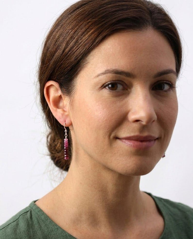 May include: A drop earring featuring a vertical row of small, faceted beads in a gradient of pink and deep magenta hues. The beads are strung on a thin silver-toned wire with a simple hook closure, creating a delicate and slender dangling design.