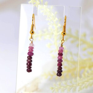 May include: A pair of drop earrings featuring gold-toned fishhook wires and a vertical strand of faceted gemstone beads in a gradient of pink and deep red hues, ranging from light pale pink at the top to dark burgundy at the bottom.