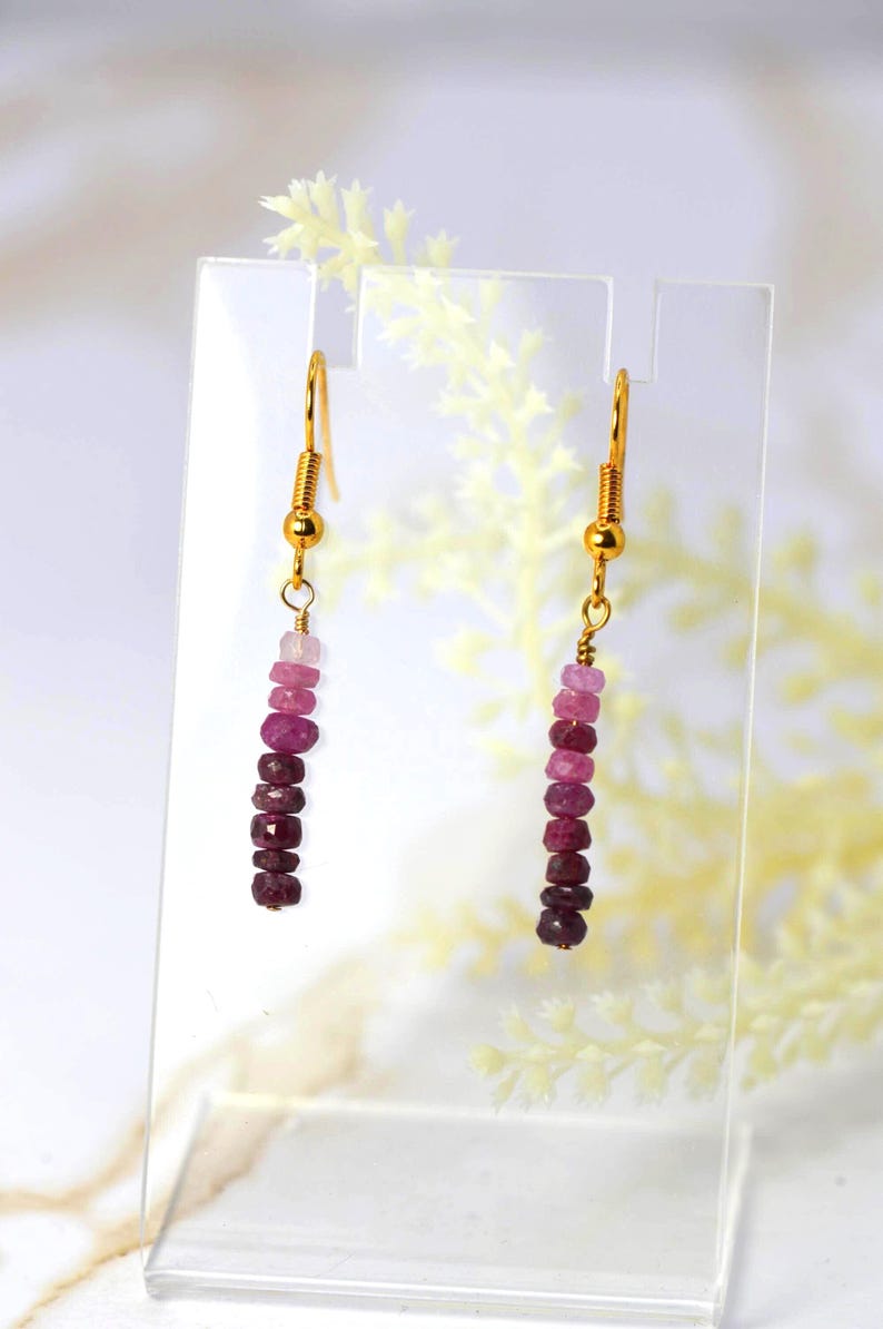 May include: A pair of drop earrings featuring gold-toned fishhook wires and a vertical strand of faceted gemstone beads in a gradient of pink and deep red hues, ranging from light pale pink at the top to dark burgundy at the bottom.