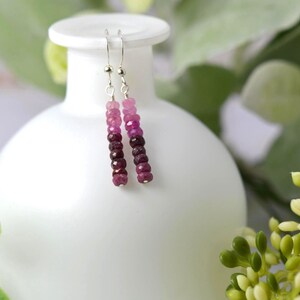 May include: A pair of drop earrings featuring a vertical row of faceted gemstone beads in an ombre colour gradient, transitioning from light pink at the top to deep magenta and dark burgundy at the bottom, suspended from silver-toned metal hooks.