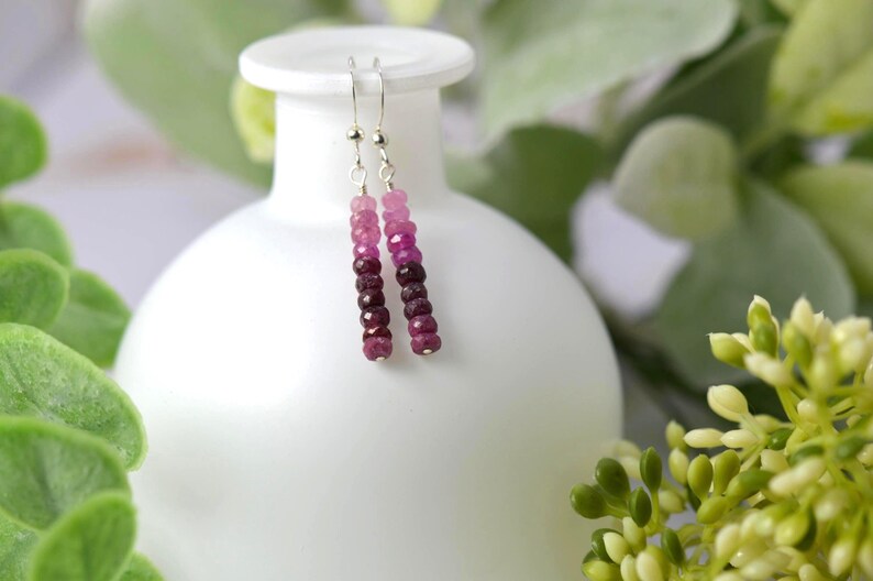 May include: A pair of drop earrings featuring a vertical row of faceted gemstone beads in an ombre colour gradient, transitioning from light pink at the top to deep magenta and dark burgundy at the bottom, suspended from silver-toned metal hooks.