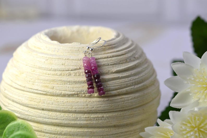 May include: A pair of drop earrings featuring small, faceted, cylindrical beads in varying shades of pink and deep magenta. The beads are strung on thin silver-toned wire with standard hook-style ear wires for attachment to the earlobe.