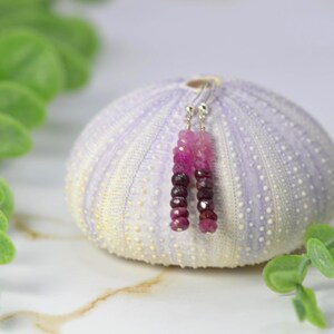 May include: A pair of dangle earrings featuring faceted gemstone beads in a gradient of pink to deep burgundy. The beads are strung on thin silver-toned metal wires with standard hook-style ear wires for attachment, creating a vertical drop design.