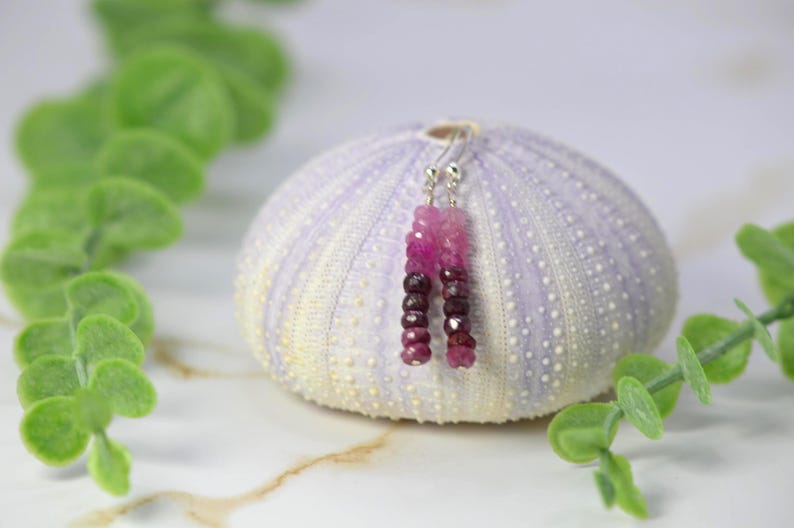May include: A pair of dangle earrings featuring faceted gemstone beads in a gradient of pink to deep burgundy. The beads are strung on thin silver-toned metal wires with standard hook-style ear wires for attachment, creating a vertical drop design.