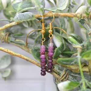 May include: A pair of dangle earrings featuring gold-toned metal hooks and a vertical arrangement of small, faceted, round beads in a gradient of pink and deep magenta hues. The beads are strung on thin wire, creating a slender, linear drop design.