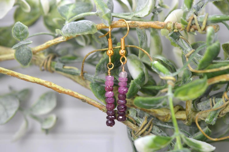 May include: A pair of dangle earrings featuring gold-toned metal hooks and a vertical arrangement of small, faceted, round beads in a gradient of pink and deep magenta hues. The beads are strung on thin wire, creating a slender, linear drop design.