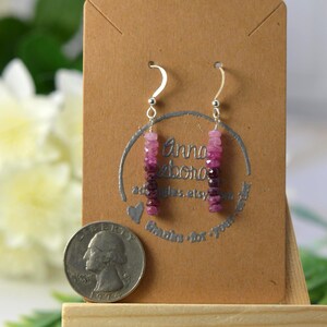 May include: Dangle earrings featuring faceted pink and deep magenta gemstone beads on silver-toned hooks. The earrings are displayed on a brown card next to a United States quarter coin for scale, showing the beads arranged in a vertical gradient.