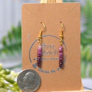 May include: A pair of dangle earrings featuring a vertical row of small, faceted beads in a gradient of pink to deep purple tones. The earrings are suspended from gold-toned metal hooks and are displayed on a brown card next to a US quarter coin.