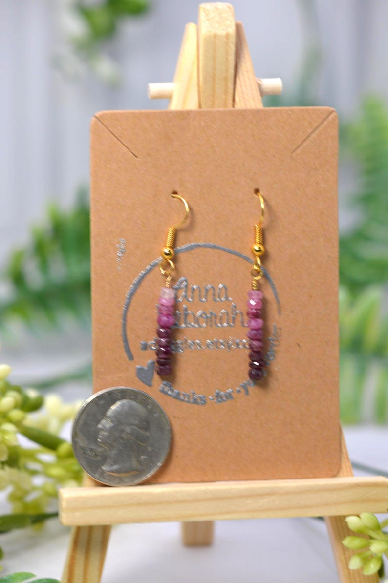 May include: A pair of dangle earrings featuring a vertical row of small, faceted beads in a gradient of pink to deep purple tones. The earrings are suspended from gold-toned metal hooks and are displayed on a brown card next to a US quarter coin.